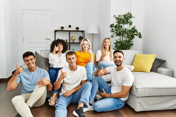 Group of people sitting on the sofa and floor at home smiling happy and positive, thumb up doing excellent and approval sign