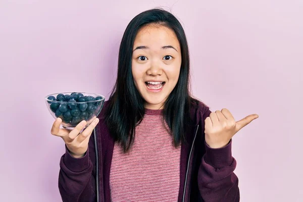 Young Chinese Girl Holding Blueberries Pointing Thumb Side Smiling Happy — Φωτογραφία Αρχείου