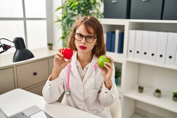 Young Doctor Woman Holding Heart Green Apple Skeptic Nervous Frowning — 图库照片