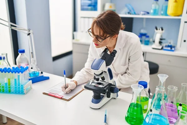 Young Caucasian Woman Wearing Scientist Uniform Writing Clipboard Using Microscope — стоковое фото
