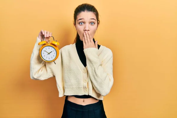 Young Brunette Girl Holding Alarm Clock Covering Mouth Hand Shocked — 图库照片