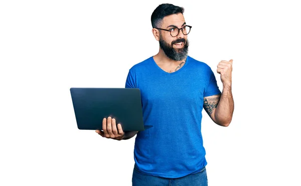 一个留着胡子的西班牙男人用电脑笔记本电脑把大拇指指向旁边 笑着张开了嘴 — 图库照片