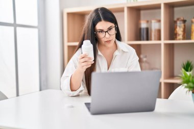 Young hispanic woman holding pills having telemedicine at home clipart