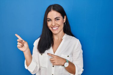Mavi arka planda dikilen genç İspanyol kadın gülümsüyor ve kameraya bakıyor iki eliyle ve parmaklarıyla kenarı işaret ediyor.. 