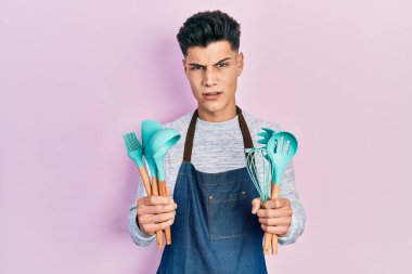 Young hispanic man wearing professional baker apron holding cooking tools in shock face, looking skeptical and sarcastic, surprised with open mouth 