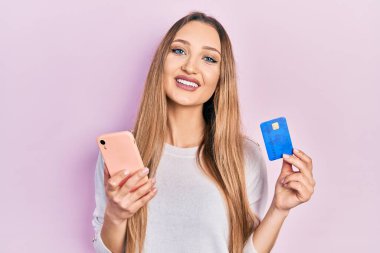 Akıllı telefon ve kredi kartı tutan genç sarışın kız mutlu ve havalı bir gülümsemeyle gülümsüyor. diş gösterme. 