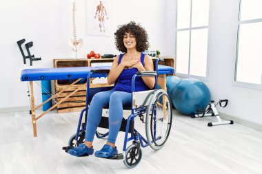 Young middle eastern woman sitting on wheelchair at physiotherapy clinic smiling with hands on chest with closed eyes and grateful gesture on face. health concept. 