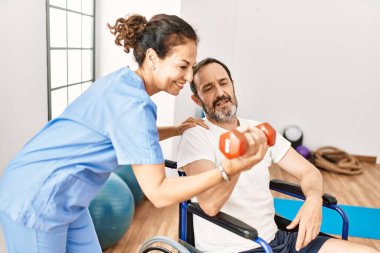 Orta yaşlı bir adam ve kadın, fizyoterapi kliniğinde tekerlekli sandalyede oturan dambılı kullanarak rehabilitasyon seansı yapıyorlar.