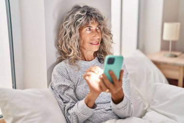 Orta yaşlı bir kadın yatakta akıllı telefon kullanıyor.