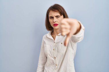 Young beautiful woman standing casual over blue background looking unhappy and angry showing rejection and negative with thumbs down gesture. bad expression. 
