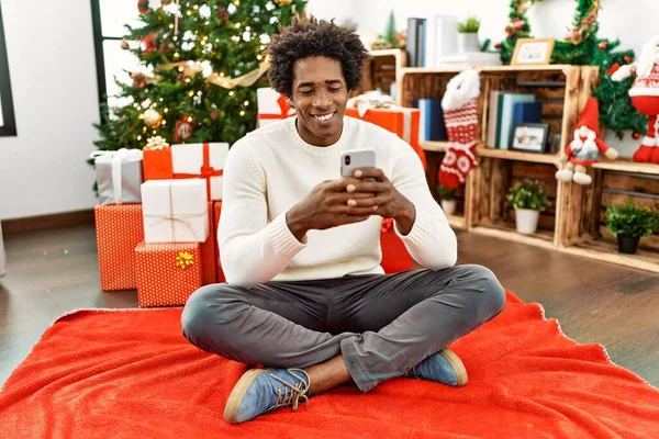 若いアフリカ系アメリカ人の男を使用してスマートフォンを床に座ってクリスマスツリーによって自宅 — ストック写真