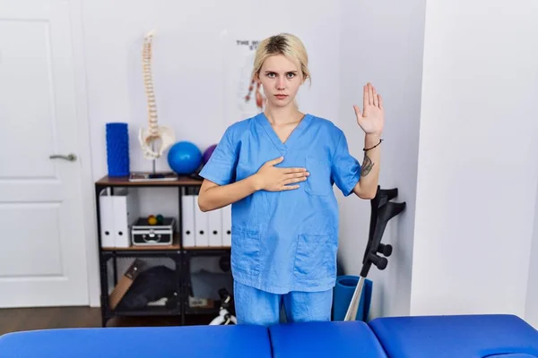 Young physiotherapist woman working at pain recovery clinic swearing with hand on chest and open palm, making a loyalty promise oath