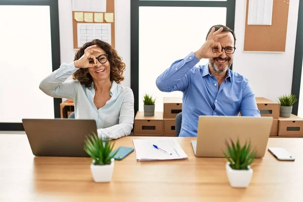 Middle age hispanic woman and man sitting with laptop at the office doing ok gesture with hand smiling, eye looking through fingers with happy face.