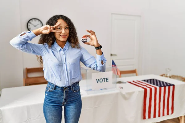 Beautiful hispanic woman standing by at political campaign by voting ballot doing peace symbol with fingers over face, smiling cheerful showing victory