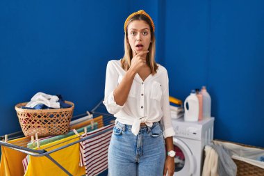 Young blonde woman at laundry room looking fascinated with disbelief, surprise and amazed expression with hands on chin 