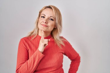 Blonde woman standing over isolated background thinking concentrated about doubt with finger on chin and looking up wondering 