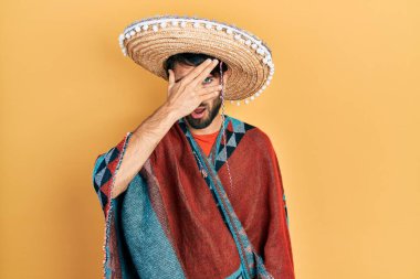 Young hispanic man holding mexican hat peeking in shock covering face and eyes with hand, looking through fingers with embarrassed expression. 