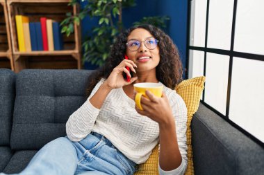Genç Latin kadın kahve içiyor ve evde akıllı telefondan konuşuyor.