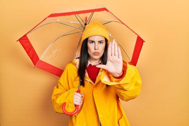 Young hispanic woman wearing yellow raincoat holding umbrella with open hand doing stop sign with serious and confident expression, defense gesture 