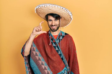 Young hispanic man holding mexican hat smiling and confident gesturing with hand doing small size sign with fingers looking and the camera. measure concept. 
