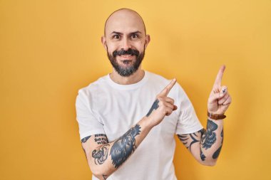 Young hispanic man with tattoos standing over yellow background smiling and looking at the camera pointing with two hands and fingers to the side. 