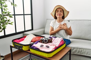 Beautiful middle age hispanic woman packing summer clothes in suitcase smiling with hands on chest with closed eyes and grateful gesture on face. health concept. 