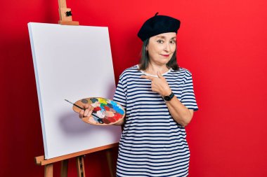 Middle age hispanic woman standing drawing with palette by painter easel stand pointing with hand finger to the side showing advertisement, serious and calm face 