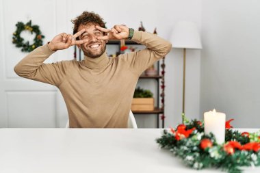 Masada sakallı genç yakışıklı adam Noel süslemeleri ile oturuyor. Barış sembolü yapıyor. Yüzünde parmaklar, gülümseyerek zaferi gösteriyor. 