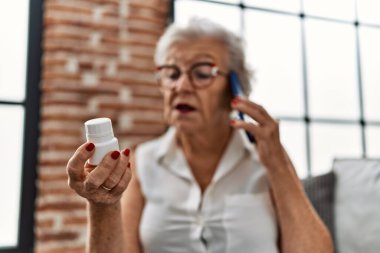 Yaşlı gri saçlı kadın evde elinde haplarla akıllı telefondan konuşuyor.