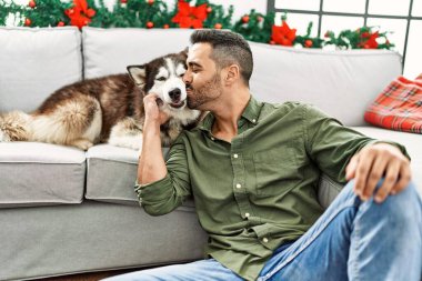 Young hispanic man kissing dog sitting on sofa by christmas decor at home