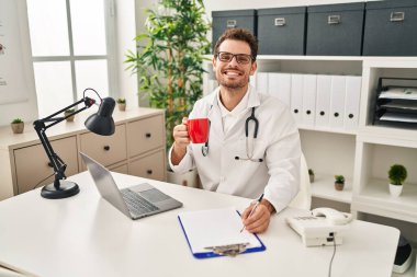Doktor üniforması giyen, kahve içen genç İspanyol adam klinikte çalışıyor.