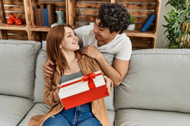 Mutlu gülümseyen genç çift evde yıldönümü hediyesiyle koltukta oturuyor..