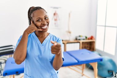 Black woman with braids working at pain recovery clinic smiling doing talking on the telephone gesture and pointing to you. call me. 