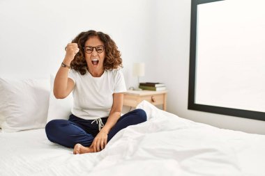 Middle age hispanic woman sitting on the bed at home angry and mad raising fist frustrated and furious while shouting with anger. rage and aggressive concept. 