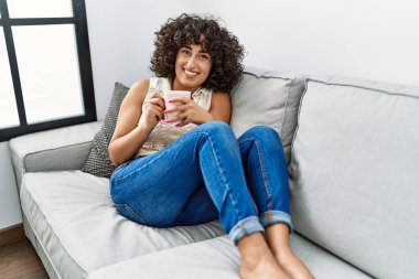 Orta Doğulu genç bir kadın gülümseyerek evde kahve içiyor.