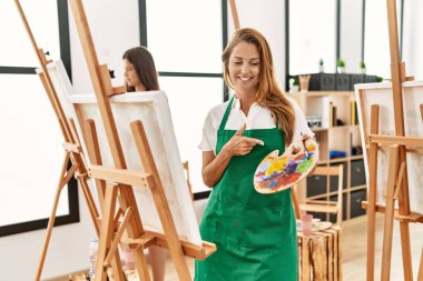 Young hispanic middle age woman at art classroom smiling happy pointing with hand and finger 