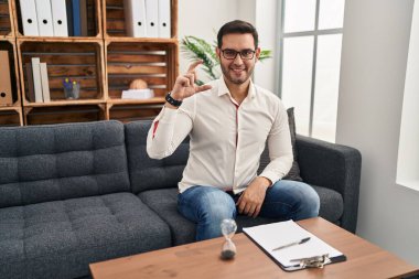 Young hispanic man with beard working at consultation office smiling and confident gesturing with hand doing small size sign with fingers looking and the camera. measure concept. 