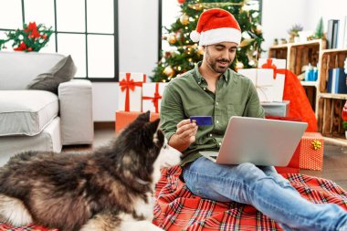 Genç İspanyol adam dizüstü bilgisayar ve kredi kartı kullanıyor evdeki yılbaşı ağacında köpekle birlikte yerde oturuyor.