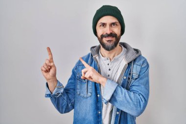 Young hispanic man with tattoos wearing wool cap smiling and looking at the camera pointing with two hands and fingers to the side. 