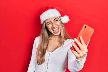 Güzel İspanyol kadın Noel şapkası takıyor akıllı telefon kullanıyor kameraya seksi bir ifadeyle bakıyor, neşeli ve mutlu bir yüzle.. 