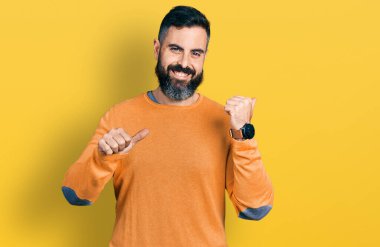 Sakallı, sıradan bir süveter giyen İspanyol bir adam. Arka tarafı işaret ediyor. El ve baş parmakları havada. 