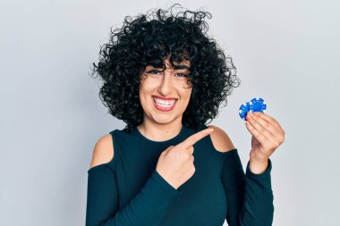 Young middle east woman playing poker holding casino chips smiling happy pointing with hand and finger 