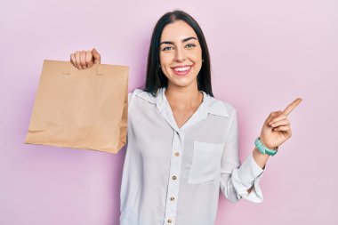 Beautiful woman with blue eyes holding take away paper bag smiling happy pointing with hand and finger to the side 