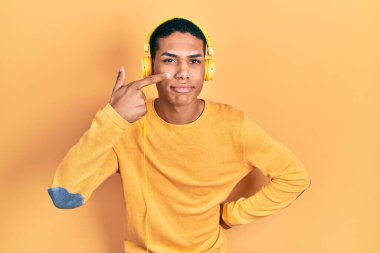 Genç Afro-Amerikalı adam müzik dinliyor kulaklıkla el ve burun parmaklarıyla işaret ediyor, neşeyle gülümsüyor. güzellik konsepti 