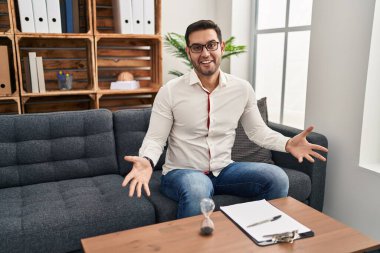 Young hispanic man with beard working at consultation office smiling cheerful with open arms as friendly welcome, positive and confident greetings 