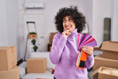 Young brunette woman with curly hair choosing color of new house wall serious face thinking about question with hand on chin, thoughtful about confusing idea 