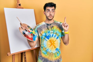 İspanyol asıllı genç adam paletle resim çiziyor, resim sehpası ayakta üzgün ve üzgün görünüyor, parmaklarıyla yönü gösteriyor, mutsuz ve depresif.. 