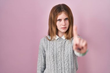Beautiful woman standing over pink background pointing with finger up and angry expression, showing no gesture 