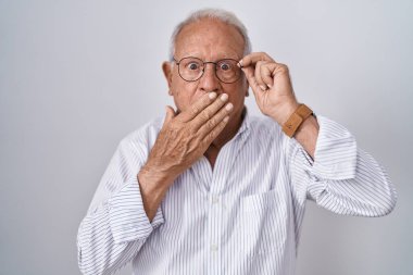 Senior man with grey hair holding glasses with hand covering mouth with hand, shocked and afraid for mistake. surprised expression 