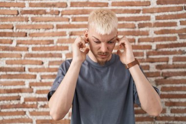 Young caucasian man standing over bricks wall covering ears with fingers with annoyed expression for the noise of loud music. deaf concept. 